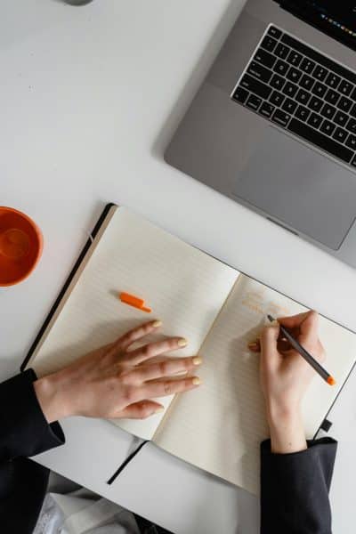 pexels-photo-7718642-7718642 Person writing notes in a notepad next to a laptop, viewed from above.